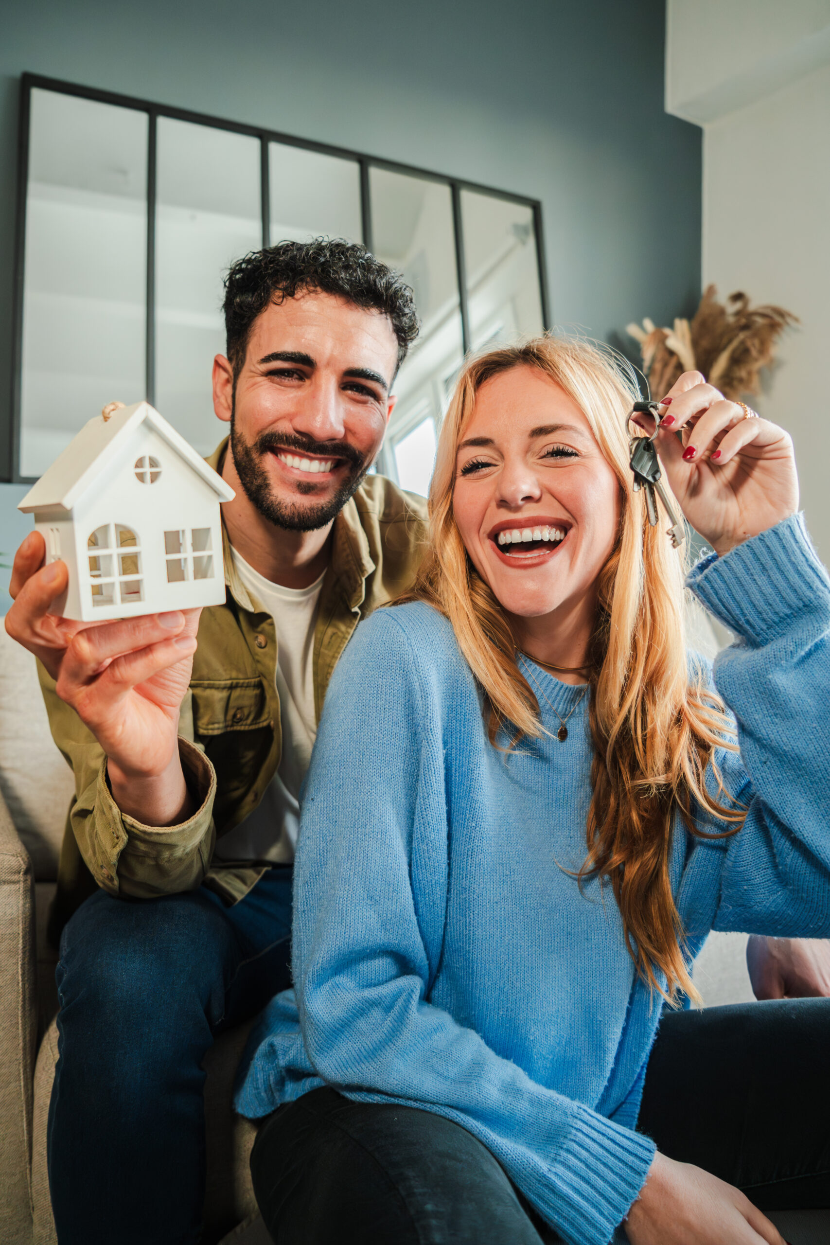 Vertical. couple celebrating a beginning at their new home holding the key of the house. Real estate investment. Mortgage or loan ad. Happy property owners at living room. Married people achievement
